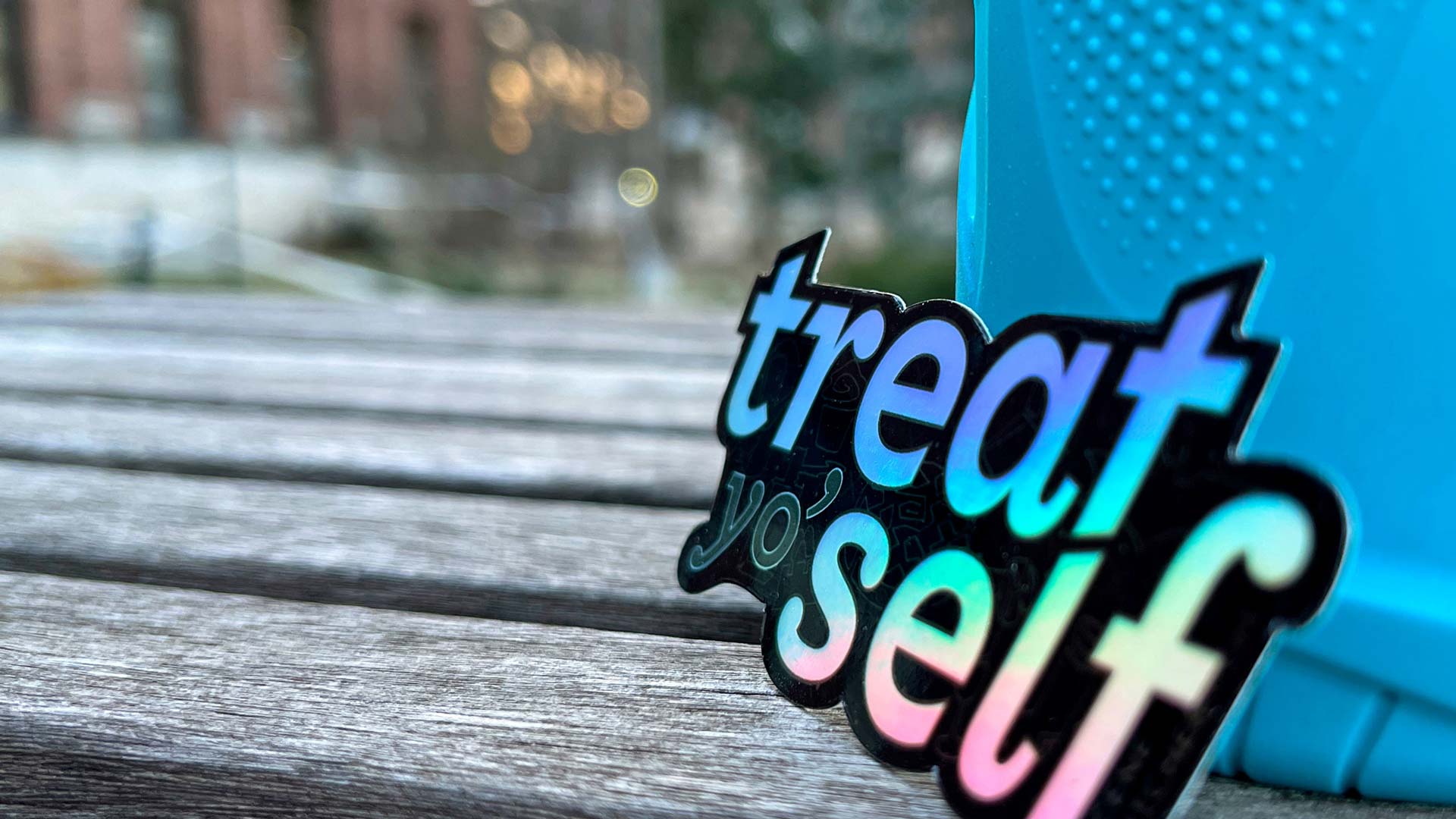 Vinyl “treat yo’ self” sticker with holographic finish propped against water bottle on outdoor table